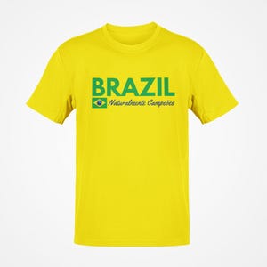 May include: A yellow t-shirt with the word "BRAZIL" in green block letters. Below the word "BRAZIL" is the Brazilian flag and the words "Naturalmente Campe&otilde;es" in green. The shirt has short sleeves and a crew neck.