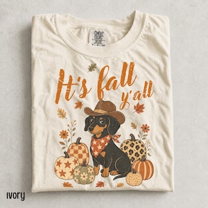 May include: An ivory t-shirt featuring a graphic of a dachshund wearing a brown cowboy hat and a red star-patterned bandana. The text reads "It's fall y'all" in orange, surrounded by illustrations of pumpkins with various patterns and autumn leaves.