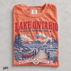 K&ouml;nnte beinhalten: Ein korallenfarbenes T-Shirt mit einer Grafik des Lake Ontario, die ein rotes Kanu, einen Holzsteg, einen Leuchtturm und eine Skyline zeigt. Der Text lautet LAKE ONTARIO ONTARIO'S FRESHWATER COAST und GREAT LAKES CANADA mit einem Anker.