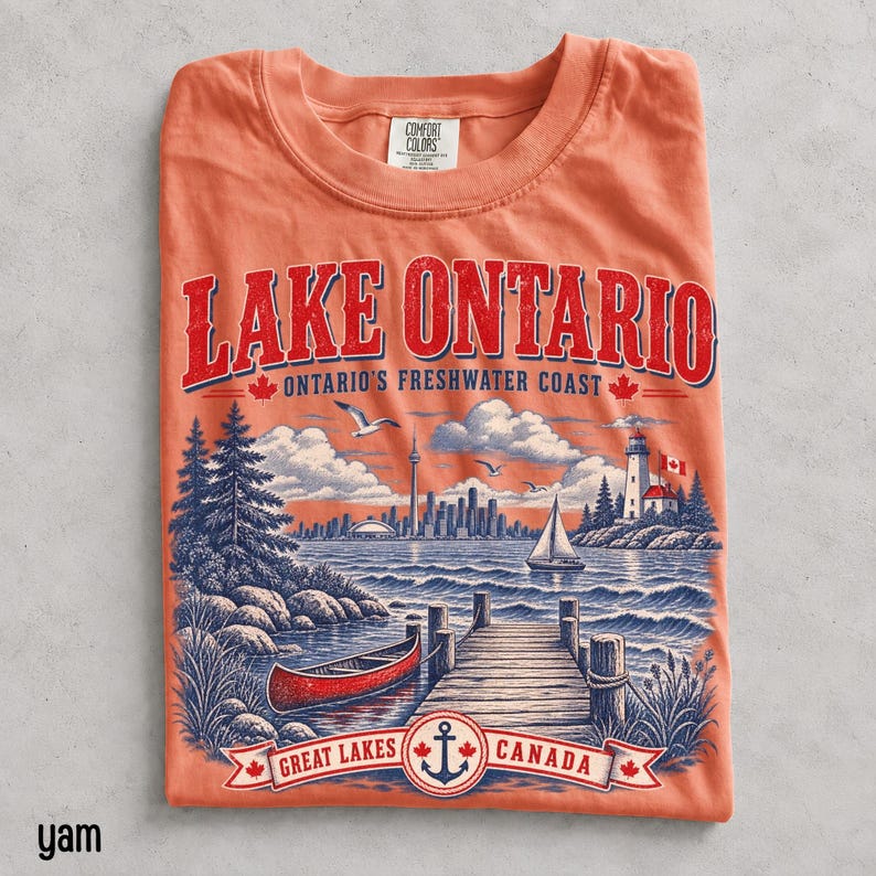 K&ouml;nnte beinhalten: Ein korallenfarbenes T-Shirt mit einer Grafik des Lake Ontario, die ein rotes Kanu, einen Holzsteg, einen Leuchtturm und eine Skyline zeigt. Der Text lautet LAKE ONTARIO ONTARIO'S FRESHWATER COAST und GREAT LAKES CANADA mit einem Anker.