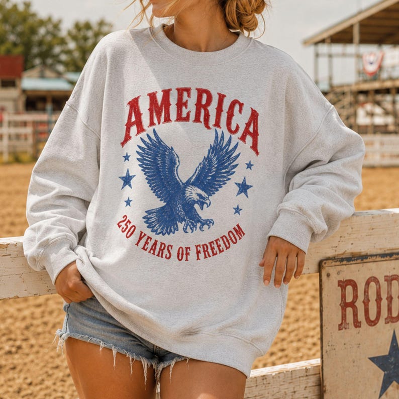 May include: A light gray sweatshirt with the word "AMERICA" in red, arched above a blue eagle graphic. Below the eagle, the text reads "250 YEARS OF FREEDOM" in red. The sweatshirt is a casual style.