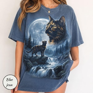 May include: A blue short-sleeved t-shirt featuring a graphic print of a large cat head in the sky above a smaller cat standing on a rock near a waterfall, a forest, and a full moon. The shirt has a faded, washed-out appearance and a crew neckline.