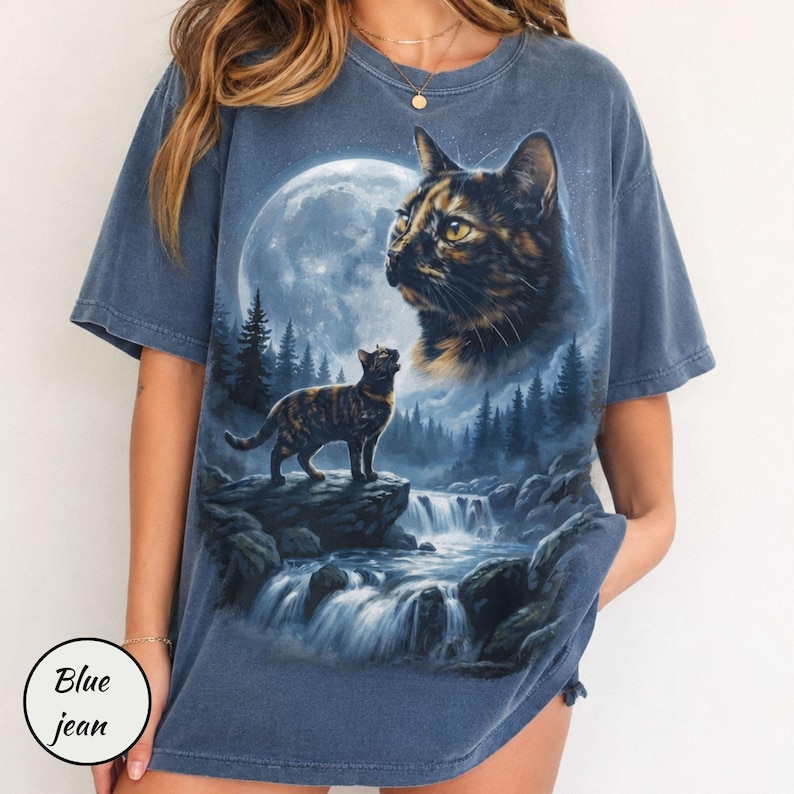 May include: A blue short-sleeved t-shirt featuring a graphic print of a large cat head in the sky above a smaller cat standing on a rock near a waterfall, a forest, and a full moon. The shirt has a faded, washed-out appearance and a crew neckline.