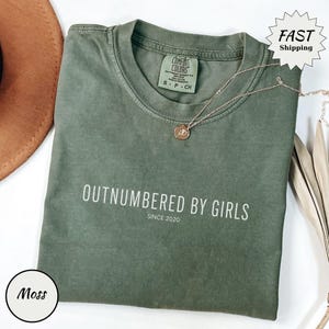 May include: A moss green t-shirt with the text "OUTNUMBERED BY GIRLS SINCE 2020" in white. A gold-colored necklace with a pendant is draped on the shirt. The shirt is folded and displayed with a brown hat and decorative leaves.