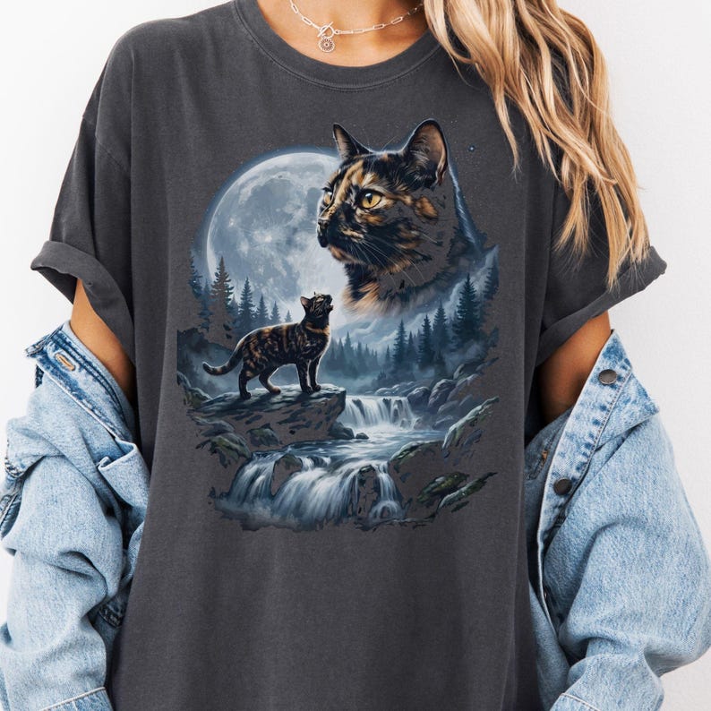 May include: A charcoal gray short-sleeve t-shirt featuring a graphic print of a tortoiseshell cat standing on a rock near a waterfall, with a large moon and a forest in the background and a larger portrait of the same cat above the scene.