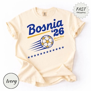 Può includere: Una t-shirt a maniche corte color avorio con testo grafico blu e giallo che recita Bosnia '26 Soccer Club. Il design include un pallone da calcio con stelle gialle, linee di movimento e una fila orizzontale di undici stelle blu sotto la grafica.