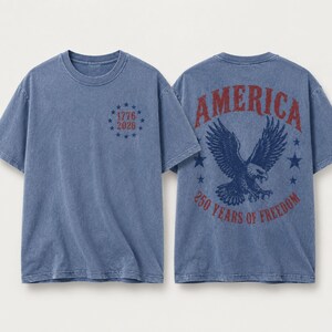 May include: A blue short-sleeve t-shirt featuring a small circle of stars with the text 1776 2026 on the front. The back displays a large eagle graphic with the text AMERICA 250 YEARS OF FREEDOM in red and blue, set against a vintage-wash fabric.