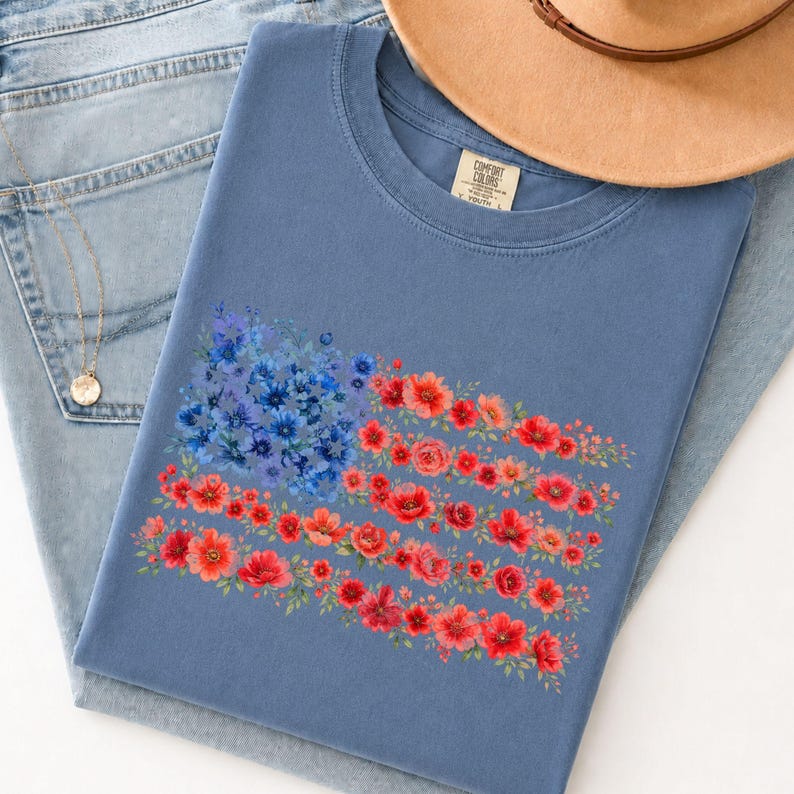 May include: A blue t-shirt featuring a floral American flag design. The flag is composed of blue and red flowers, with a tan hat and blue jeans in the background. The shirt has a tag that says "Comfort Colors".