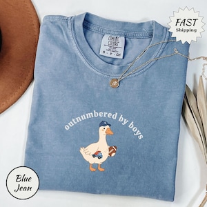 May include: Blue jean Comfort Colors t-shirt with the phrase "outnumbered by boys" and a cartoon duck wearing a baseball cap and holding a football. The duck also has a toy car. A gold necklace is draped across the shirt.