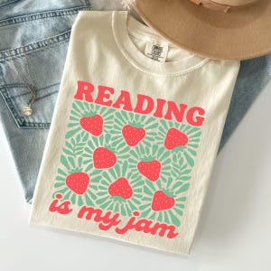 May include: Cream-colored t-shirt with the text "READING is my jam" in red, with a strawberry and leaf design. The shirt is folded, with a pair of blue jeans and a tan hat in the background.