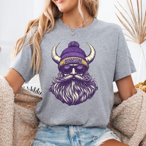Pu&ograve; includere: T-shirt grigia con una grafica di un uomo barbuto con un berretto viola con "MINNESOTA" e occhiali da sole. L'uomo ha grandi corna gialle e una lunga barba viola. Il design &egrave; un'illustrazione stilizzata.