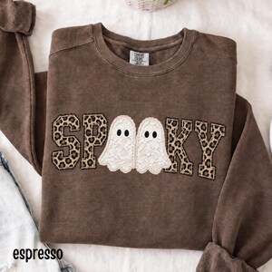 Könnte beinhalten: Ein braunes Sweatshirt mit Rundhalsausschnitt und dem Wort SPOOKY in Blockbuchstaben. Die Buchstaben S, P, O, K und Y haben ein Leopardenmuster, während die beiden O durch weiße Spitzengeister mit schwarzen Augen ersetzt sind. Comfort Colors Etikett.