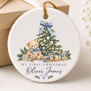 Peut inclure: Une décoration ronde en céramique blanche ornée d'un sapin de Noël, d'un ours en peluche avec un nœud à carreaux bleus et d'un train miniature. Le texte indique MY FIRST CHRISTMAS, Oliver James et 2026. Un cordon doré est fixé au sommet.