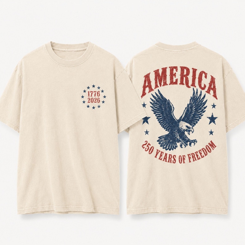 May include: A beige short-sleeve t-shirt featuring a front graphic of the text 1776 2026 surrounded by a circle of blue stars. The back displays a large blue eagle graphic with the red text AMERICA and 250 YEARS OF FREEDOM with blue stars.