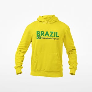 May include: A yellow hooded sweatshirt with the word "BRAZIL" in green block letters. Below the word "BRAZIL" is the Brazilian flag and the words "Naturalmente Campe&otilde;es" in green. The sweatshirt has a front pocket and long sleeves.