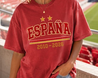 Może przedstawiać: Czerwony t-shirt z krótkim rękawem z napisem ESPAÑA dużymi czerwonymi literami ze złotym obrysem. Nad napisem znajdują się dwie złote gwiazdki. Pod napisem dwie poziome linie w kolorze czerwonym i złotym oraz złoty napis 2010-2026.