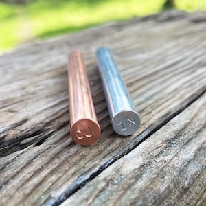 Op de afbeelding: Twee cilindrische metalen staven, één van koper met "CU" gestempeld op het uiteinde, en één zilverkleurig met "ZN" gestempeld. De staven liggen op een verweerd houten oppervlak.