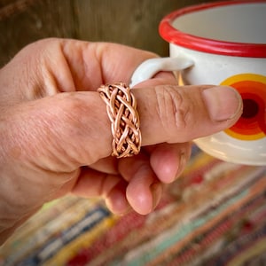 Op de afbeelding: Een koperen ring met een geweven, gevlochten metaalontwerp. De ring wordt gedragen aan een vinger naast een witte keramische mok met een rode rand en een geel en oranje cirkelvormig grafisch patroon op de zijkant, wat de metalen textuur benadrukt.