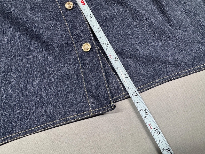 Puede incluir: Primer plano de una prenda de mezclilla azul oscuro con botones dorados y costuras blancas. Una cinta m&eacute;trica muestra una longitud de aproximadamente 53 cm.
