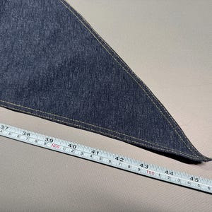 Puede incluir: Un tri&aacute;ngulo de tela vaquera azul oscuro con un borde cosido beige. Una cinta m&eacute;trica muestra que la tela mide aproximadamente 117 cm de largo. La tela est&aacute; sobre una superficie gris claro.