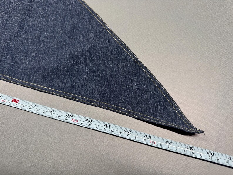 Puede incluir: Un tri&aacute;ngulo de tela vaquera azul oscuro con un borde cosido beige. Una cinta m&eacute;trica muestra que la tela mide aproximadamente 117 cm de largo. La tela est&aacute; sobre una superficie gris claro.