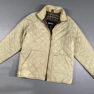 May include: A cream-colored quilted jacket with a full zipper closure and two front pockets. The jacket features a corduroy collar and a plaid lining. The brand name "Barbour" is visible on the chest. This jacket is a classic outerwear piece.