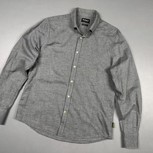 Puede incluir: Una camisa de manga larga con botones en tejido gris jaspeado. La camisa presenta un cuello clásico, una tapeta frontal con botones y puños abotonados. Una pequeña etiqueta de la marca Barbour es visible en la parte inferior izquierda, y la etiqueta interior del cuello dice Barbour.