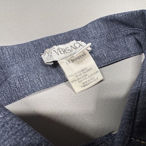 Puede incluir: Primer plano del cuello de una chaqueta vaquera azul oscuro. Una etiqueta blanca muestra el logotipo de Versace y el texto "LE BONITAS". La etiqueta tambi&eacute;n enumera la composici&oacute;n de la tela: 75% PA Poliamida, 25% EA Elastano. Hecho en Italia.