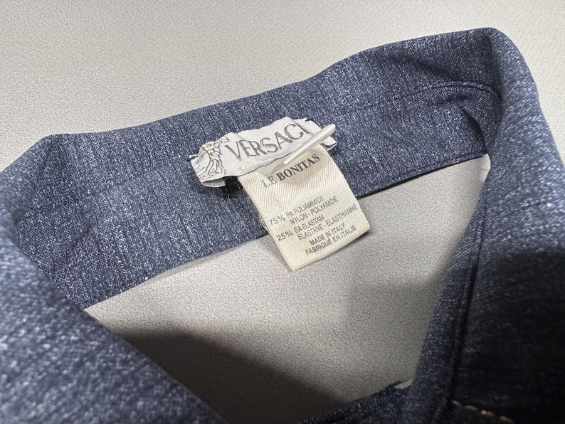 Puede incluir: Primer plano del cuello de una chaqueta vaquera azul oscuro. Una etiqueta blanca muestra el logotipo de Versace y el texto "LE BONITAS". La etiqueta tambi&eacute;n enumera la composici&oacute;n de la tela: 75% PA Poliamida, 25% EA Elastano. Hecho en Italia.