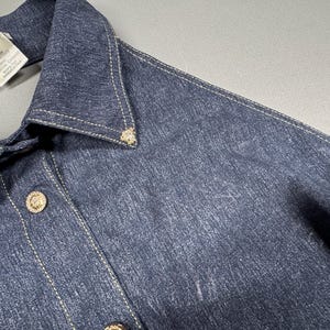 Puede incluir: Primer plano de una camisa vaquera azul oscuro con cuello y cierre de botones. La camisa presenta botones dorados y costuras decorativas. Una etiqueta con la marca "Versace" es visible cerca del cuello.