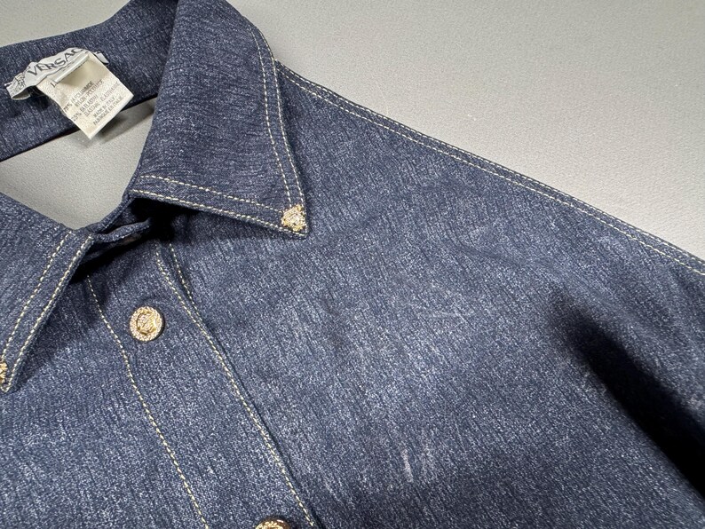 Puede incluir: Primer plano de una camisa vaquera azul oscuro con cuello y cierre de botones. La camisa presenta botones dorados y costuras decorativas. Una etiqueta con la marca "Versace" es visible cerca del cuello.