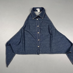 Puede incluir: Una camisa vaquera azul oscuro con un dise&ntilde;o asim&eacute;trico &uacute;nico. La camisa presenta un cuello, una parte delantera abotonada con botones dorados y mangas largas y puntiagudas. La prenda est&aacute; tendida sobre una superficie gris.