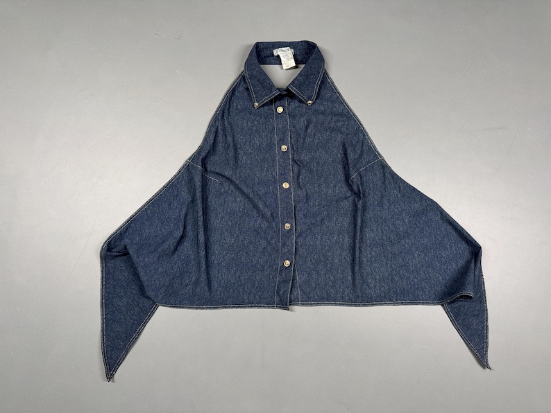 Puede incluir: Una camisa vaquera azul oscuro con un dise&ntilde;o asim&eacute;trico &uacute;nico. La camisa presenta un cuello, una parte delantera abotonada con botones dorados y mangas largas y puntiagudas. La prenda est&aacute; tendida sobre una superficie gris.