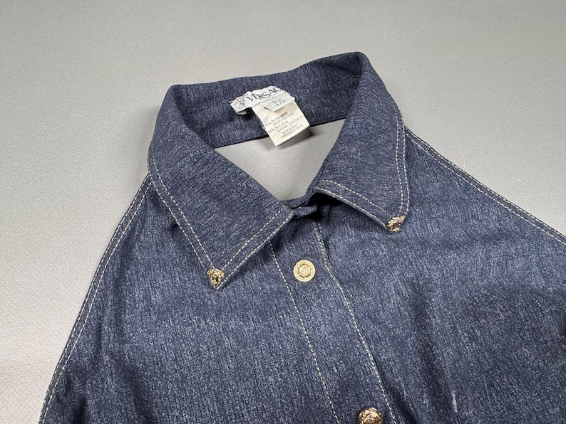 Puede incluir: Una camisa vaquera azul oscuro con cuello cl&aacute;sico y botones dorados. La camisa presenta costuras blancas a lo largo de las costuras y el cuello. La etiqueta dice "Vimsal".