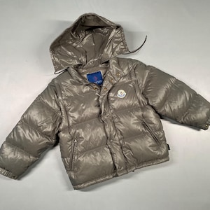 Puede incluir: Una chaqueta acolchada gris con capucha. La chaqueta tiene un diseño acolchado brillante y presenta un parche con el logotipo en el pecho. Tiene cierre de cremallera y bolsillos. La chaqueta está diseñada para niños.