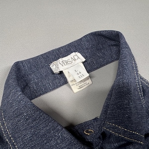 Puede incluir: Primer plano de un cuello de camisa vaquera azul oscuro con costuras blancas. La etiqueta Versace es visible en la parte superior, junto con informaci&oacute;n sobre la talla. La camisa tiene cierre de bot&oacute;n.