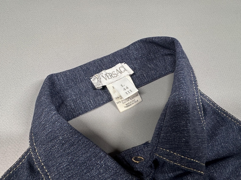 Puede incluir: Primer plano de un cuello de camisa vaquera azul oscuro con costuras blancas. La etiqueta Versace es visible en la parte superior, junto con informaci&oacute;n sobre la talla. La camisa tiene cierre de bot&oacute;n.