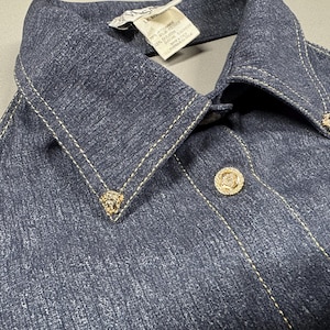 Puede incluir: Primer plano de una chaqueta vaquera azul oscuro con cuello puntiagudo y botones decorativos dorados. La chaqueta presenta costuras doradas y peque&ntilde;os adornos dorados en el cuello.