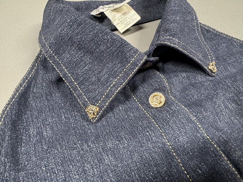 Puede incluir: Primer plano de una chaqueta vaquera azul oscuro con cuello puntiagudo y botones decorativos dorados. La chaqueta presenta costuras doradas y peque&ntilde;os adornos dorados en el cuello.