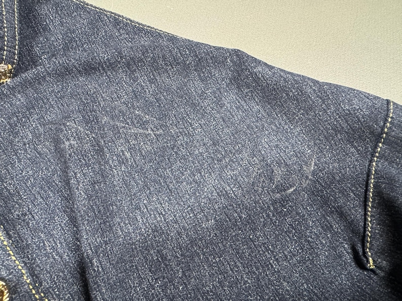 Puede incluir: Primer plano de una chaqueta vaquera azul oscuro. La tela tiene un aspecto texturizado con costuras visibles a lo largo de las costuras. El color de la chaqueta es un &iacute;ndigo profundo, con sutiles variaciones de tono.