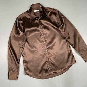 Puede incluir: Una camisa de satén marrón con cuello puntiagudo y mangas largas. La camisa tiene una textura brillante y está abotonada por delante. Los puños están abotonados. La camisa está colocada sobre una superficie gris.