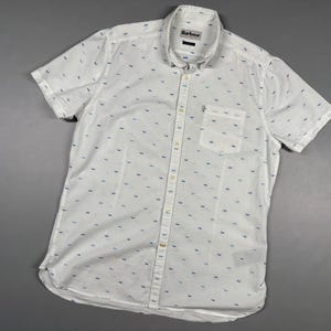 Puede incluir: Una camisa blanca de manga corta con botones y un patrón repetitivo de pequeñas formas azules y azul claro. La camisa incluye un cuello, una tapeta de botones frontal y un bolsillo en el pecho. La etiqueta de la marca Barbour es visible.