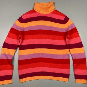 Puede incluir: Un suéter de cuello alto a rayas con tonos rojos, naranjas y rosas. El suéter tiene mangas largas y un cuello alto acanalado. El fondo es gris liso.