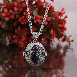 Puede incluir: Un collar con colgante plateado con un relicario ovalado. El relicario presenta un diseño ornamentado con una piedra preciosa oscura en la parte inferior y una piedra preciosa púrpura redonda más pequeña encima. La cadena del collar es de diseño de eslabones, y el fondo son flores rojas borrosas.