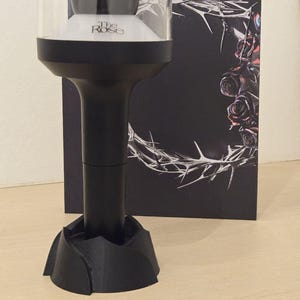 May include: A black and clear light stick with the text "The Rose" on the base. The light stick has a dome-shaped top and a black base. A black box with a thorn and rose design is in the background.