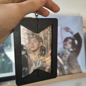 May include: Black rectangular keyring with a photo of a person inside. The keyring has a silver ring and chain. The photo shows a person with a colourful shirt. The background shows a photo of a person.
