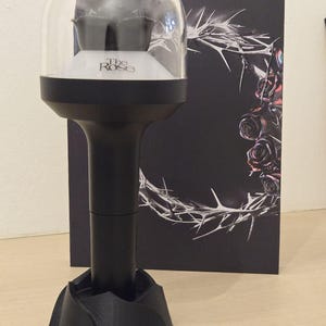May include: A black and clear light stick with the text "The Rose" on the base. The light stick has a dome-shaped top and a black base. A black box with a thorn and rose design is in the background.