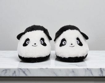 May include: A pair of plush panda slippers. Each slipper features a white body with black accents on the head, ears, and around the eyes. The slippers have embroidered facial features, including eyes and a smiling mouth. They are designed to be worn indoors.