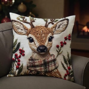 May include: A decorative square pillow featuring an embroidered deer with large eyes, antlers, and a plaid scarf. The deer is surrounded by sprigs of greenery and red berries, set against a neutral background. The pillow is placed on a gray sofa.