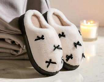 Può includere: Un paio di pantofole bianche e soffici con fiocchi neri. Le pantofole hanno una suola nera e un interno beige chiaro. Sono visibili anche una candela accesa in un portacandele e una coperta piegata.